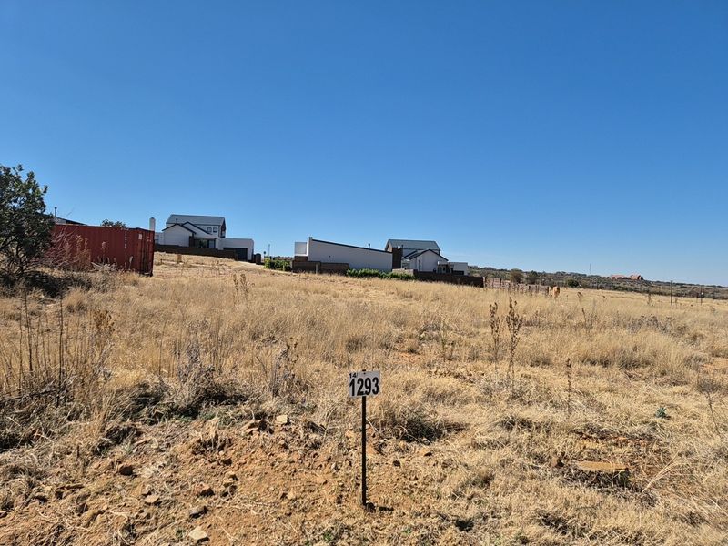 A vacant residential plot featuring dry grass and a clear property boundary marker, situated in a developing neighborhood with surrounding residential homes.
