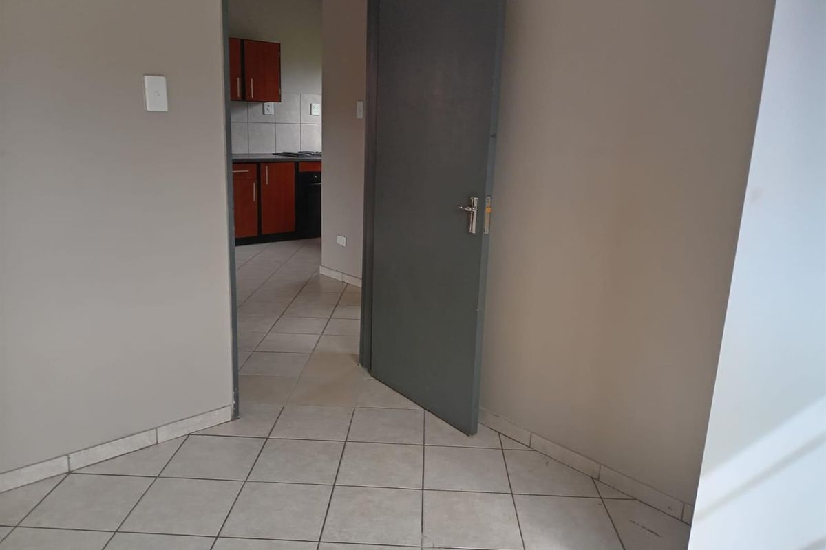 An empty living area with tiled flooring, leading through a doorway into a kitchen featuring built-in cabinetry and a hob.