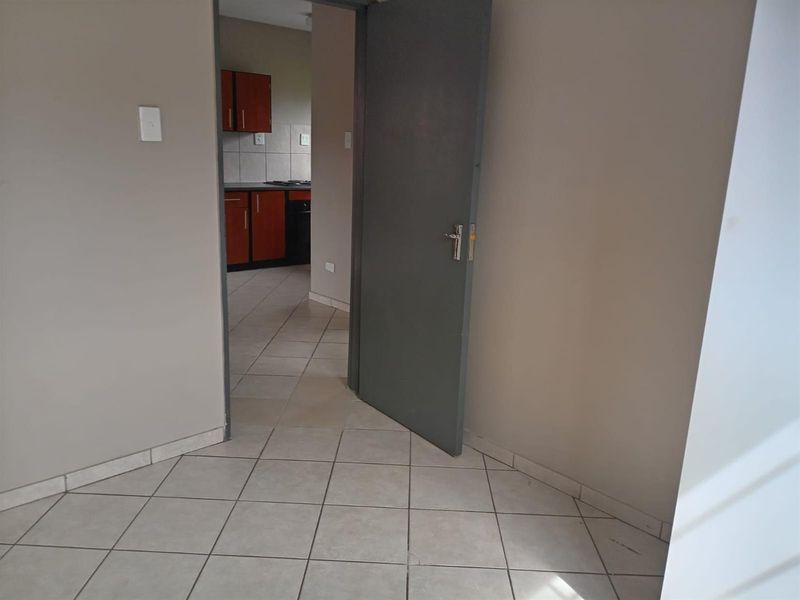 An empty living area with tiled flooring, leading through a doorway into a kitchen featuring built-in cabinetry and a hob.