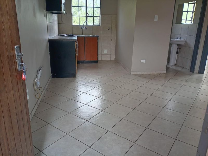 Open plan living area featuring a compact kitchen with built-in cabinetry and a separate pedestal basin area, finished with neutral floor tiles.