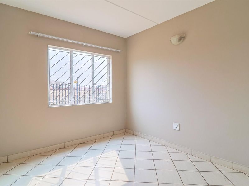 A bright, empty bedroom featuring neutral wall paint, tiled flooring, and a window fitted with security bars.