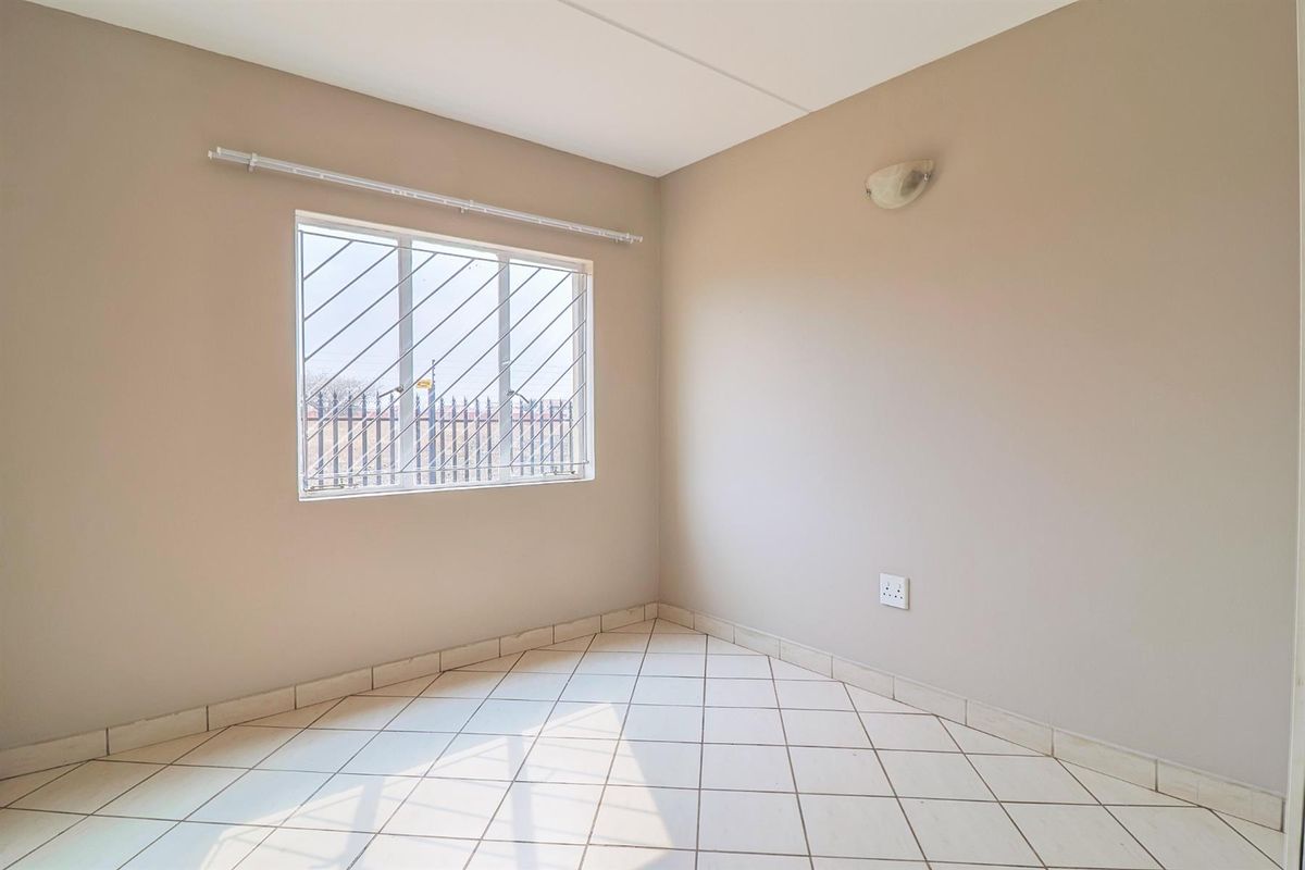A bright, empty bedroom featuring neutral wall paint, tiled flooring, and a window fitted with security bars.