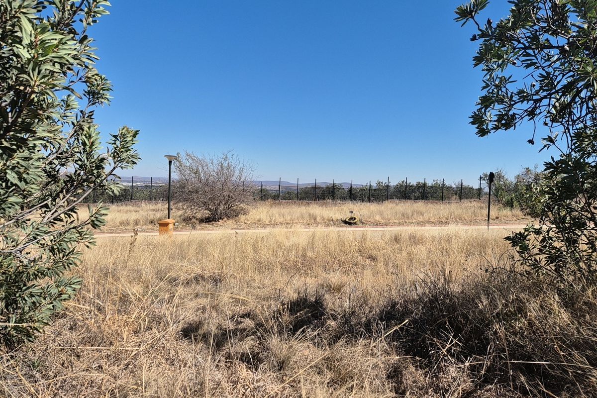 A view of a vacant residential plot featuring dry grass, perimeter fencing, and an outdoor lighting post under a clear blue sky.