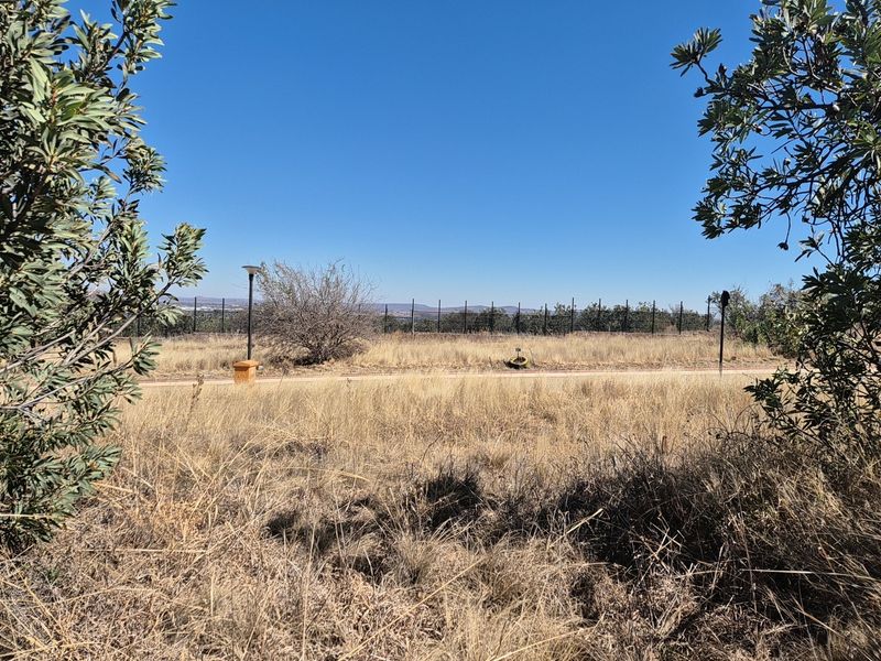 A view of a vacant residential plot featuring dry grass, perimeter fencing, and an outdoor lighting post under a clear blue sky.