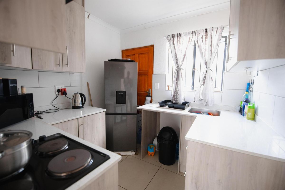 A functional kitchen featuring light wood-grain cabinetry, white countertops, a built-in electric hob, and a stainless steel sink with tiled splashbacks.