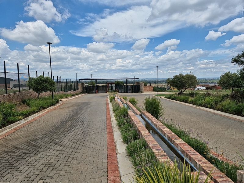 A well-maintained entrance to a residential estate featuring a paved driveway, security gate, perimeter fencing, and a decorative brick water feature.