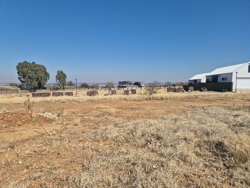 A vacant residential plot featuring dry grass, perimeter fencing, and distant views of neighboring properties under a clear blue sky.
