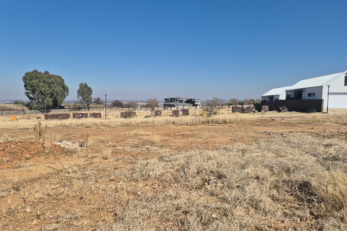 A vacant residential plot featuring dry grass, perimeter fencing, and distant views of neighboring properties under a clear blue sky.