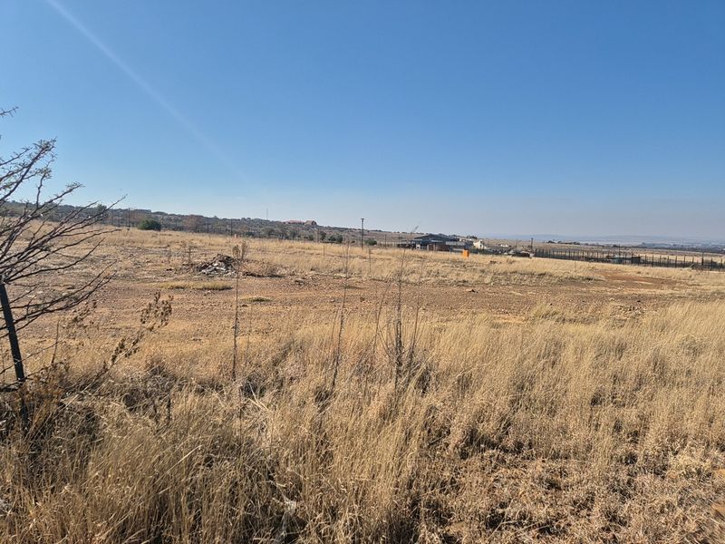 A wide-angle view of a vacant residential plot featuring dry grass and perimeter fencing under a clear blue sky.