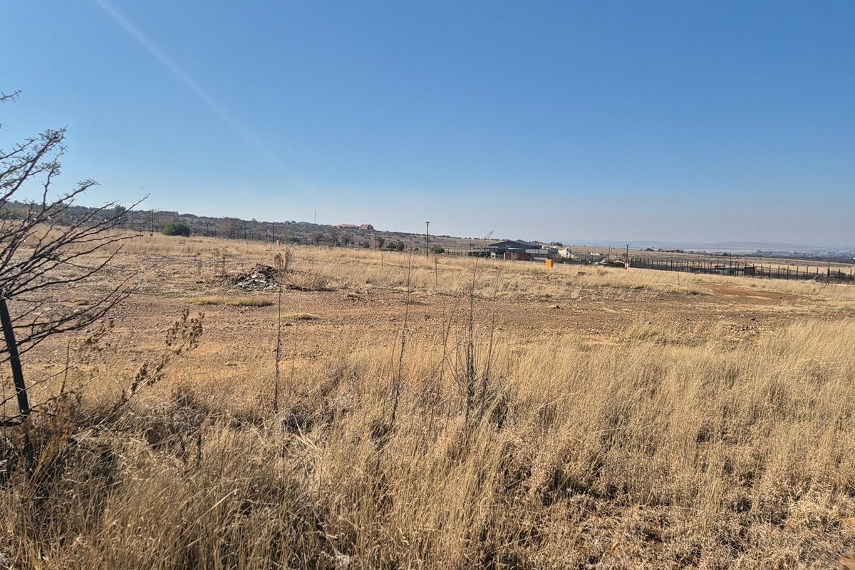 A wide-angle view of a vacant residential plot featuring dry grass and perimeter fencing under a clear blue sky.