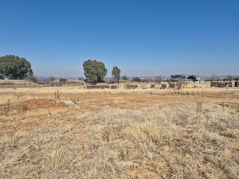 A vacant residential plot featuring dry grass, perimeter fencing, and distant views of the surrounding neighborhood.
