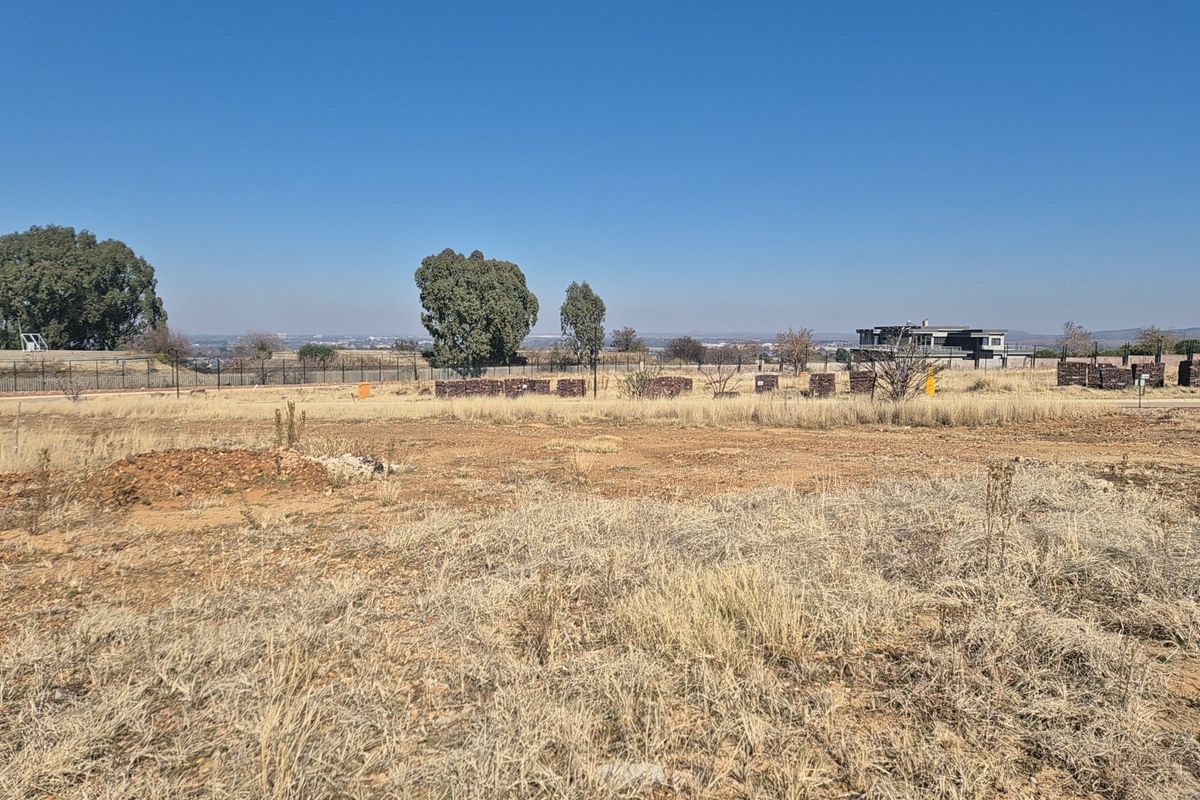 A vacant residential plot featuring dry grass, perimeter fencing, and distant views of the surrounding neighborhood.