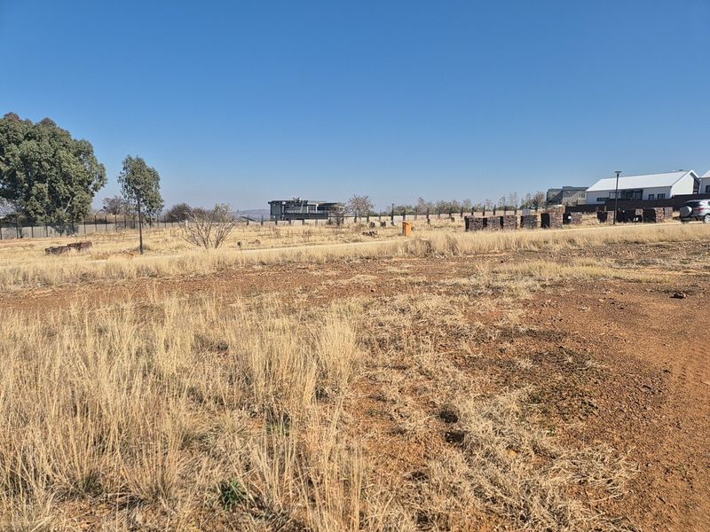 A view of a vacant residential plot featuring dry grass, perimeter fencing, and surrounding neighborhood development.