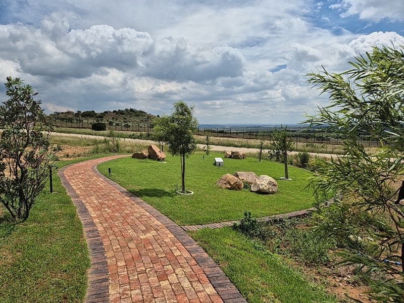 A well-maintained residential garden featuring a brick-paved pathway, manicured lawn, decorative boulders, and scenic landscape views.