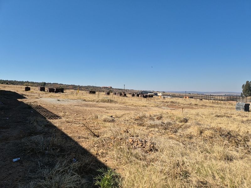 A wide, open vacant residential plot featuring dry grass, perimeter fencing, and distant landscape views under a clear blue sky.