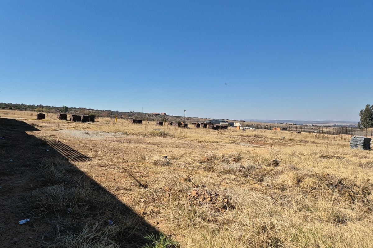 A wide, open vacant residential plot featuring dry grass, perimeter fencing, and distant landscape views under a clear blue sky.
