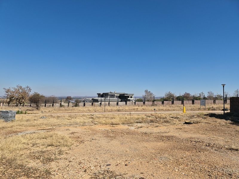 A vacant residential plot featuring dry grass, perimeter fencing, and street lighting, offering a clear view of the surrounding neighborhood.