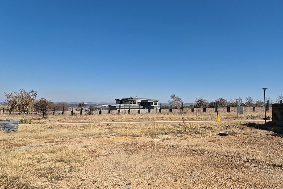 A vacant residential plot featuring dry grass, perimeter fencing, and street lighting, offering a clear view of the surrounding neighborhood.