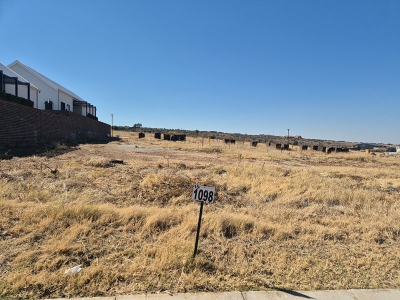 A vacant residential plot of land featuring dry grass, a boundary wall, and a clear view of the surrounding landscape.