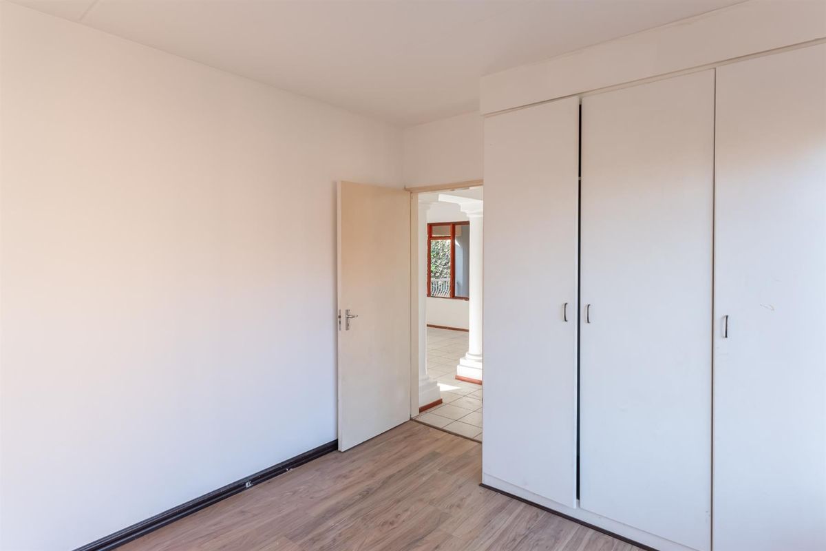 A bright, empty bedroom featuring wood-look laminate flooring and built-in white cupboards, with a doorway leading to an adjacent living area.