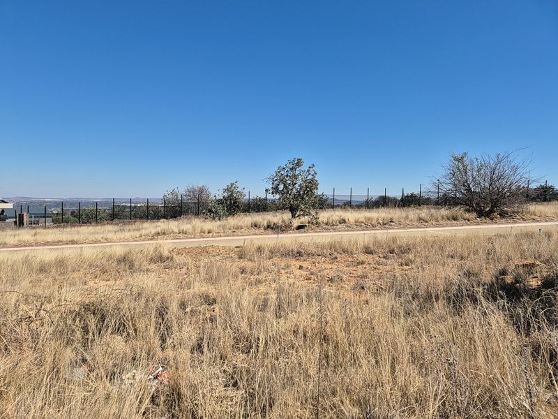 A vacant residential plot featuring natural dry grass vegetation, perimeter fencing, and direct access to a paved road.