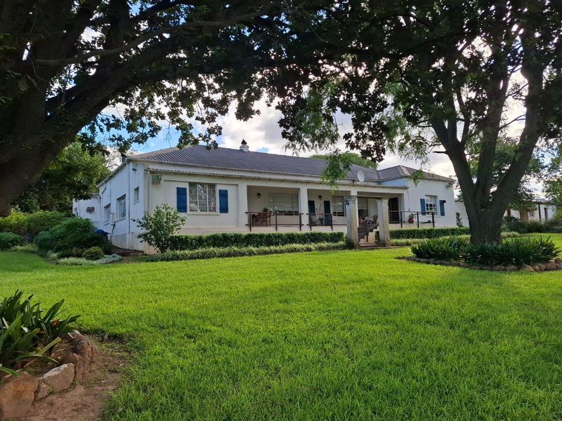 A charming farmhouse exterior featuring a covered veranda, classic blue shutters, and a well-maintained lawn surrounded by mature trees.