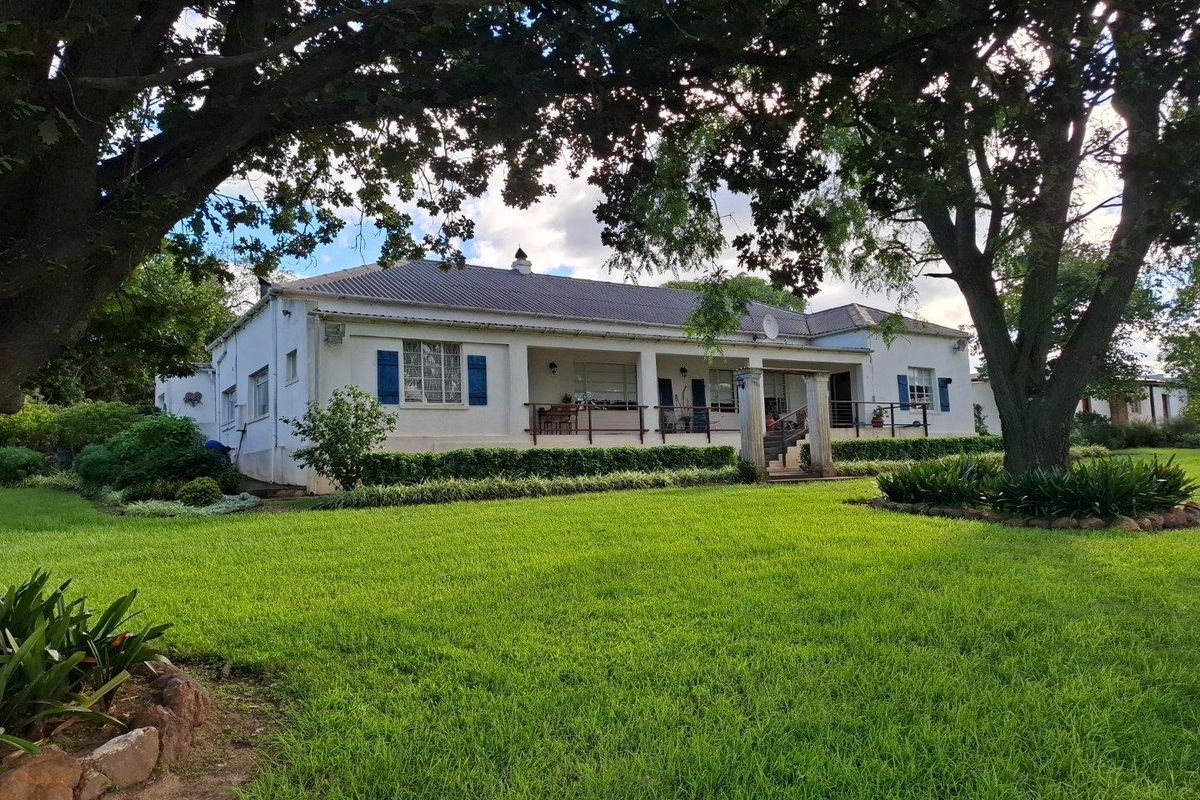 A charming farmhouse exterior featuring a covered veranda, classic blue shutters, and a well-maintained lawn surrounded by mature trees.