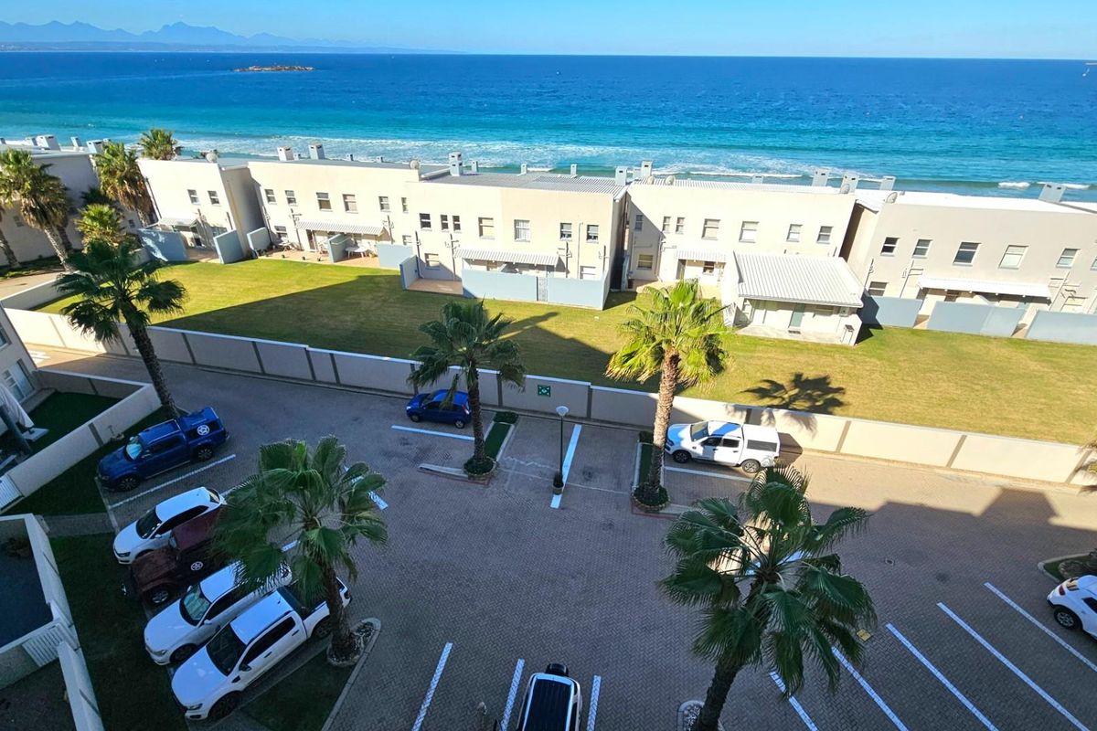 Elevated aerial view of the apartment complex grounds, featuring paved parking areas, landscaped palm trees, and direct proximity to the coastline and ocean.