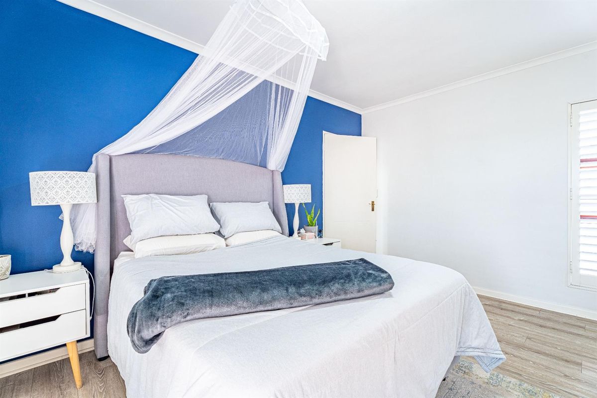 A bright bedroom featuring wood-look flooring, a bold blue accent wall, and white plantation-style shutters on the window.