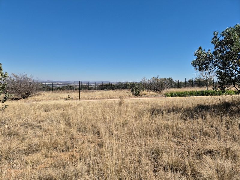 A wide-angle view of the vacant residential land, featuring natural dry grass, perimeter fencing, and distant landscape views under a clear blue sky.
