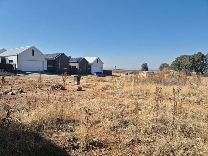 A view of a vacant residential plot of land with dry vegetation, situated adjacent to existing modern residential properties.
