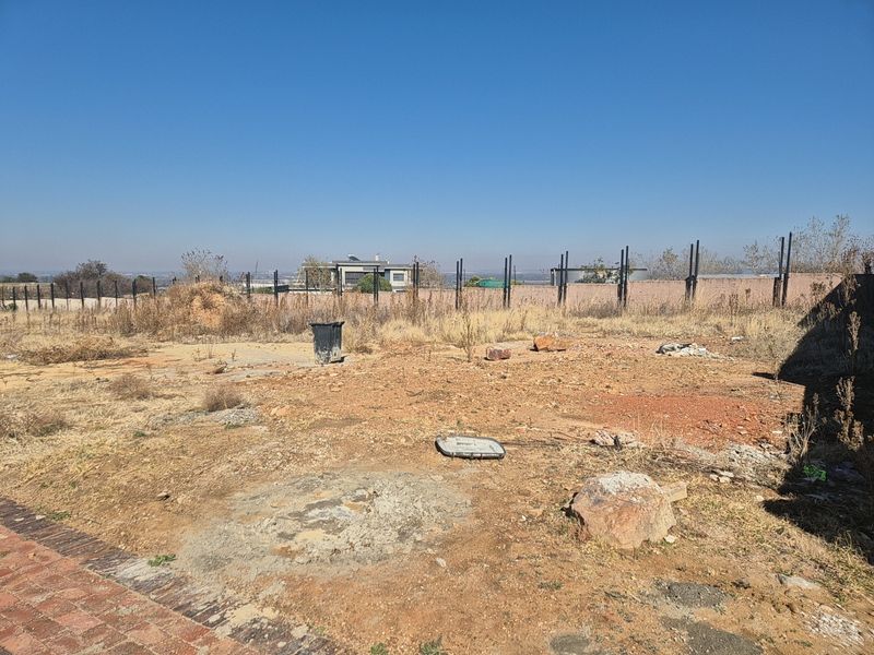 A vacant residential plot featuring dry, uneven ground, perimeter fencing, and a brick boundary wall, offering a clear view of the surrounding neighborhood.