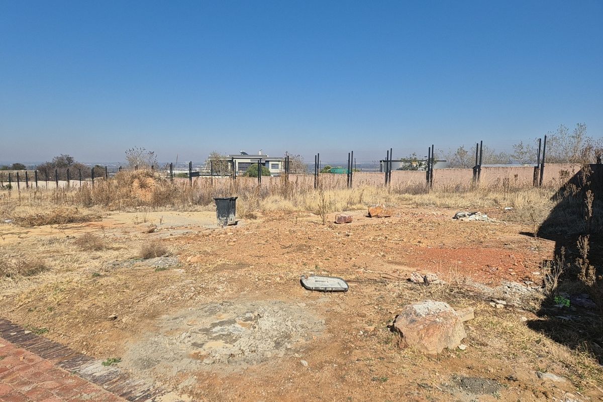 A vacant residential plot featuring dry, uneven ground, perimeter fencing, and a brick boundary wall, offering a clear view of the surrounding neighborhood.