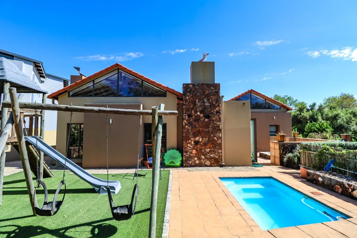 Rear view of the property featuring a swimming pool, paved deck, artificial lawn, and a distinctive stone-clad chimney with large triangular windows.