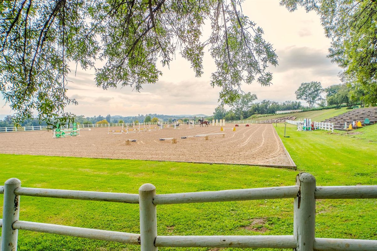 A well-maintained equestrian sports facility featuring a large sand arena with jumps, surrounded by manicured lawns and white post-and-rail fencing.