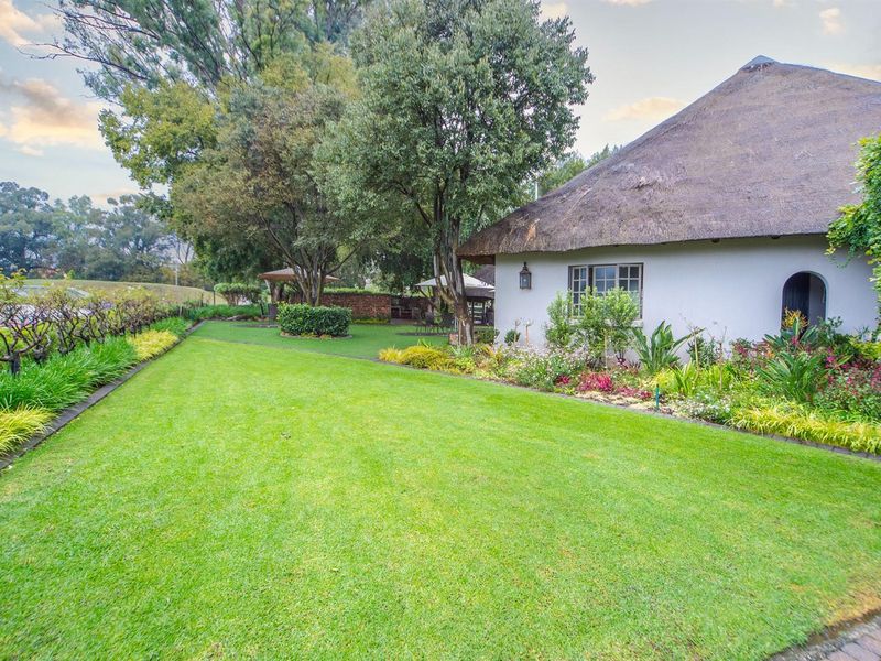 A well-maintained garden area featuring a lush green lawn, mature trees, and established flower beds adjacent to a thatched-roof residential structure.