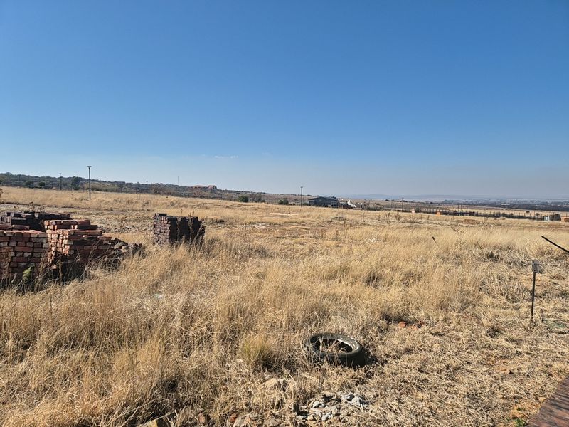 A wide-angle view of a vacant residential plot featuring dry grass, scattered building materials, and distant landscape views.