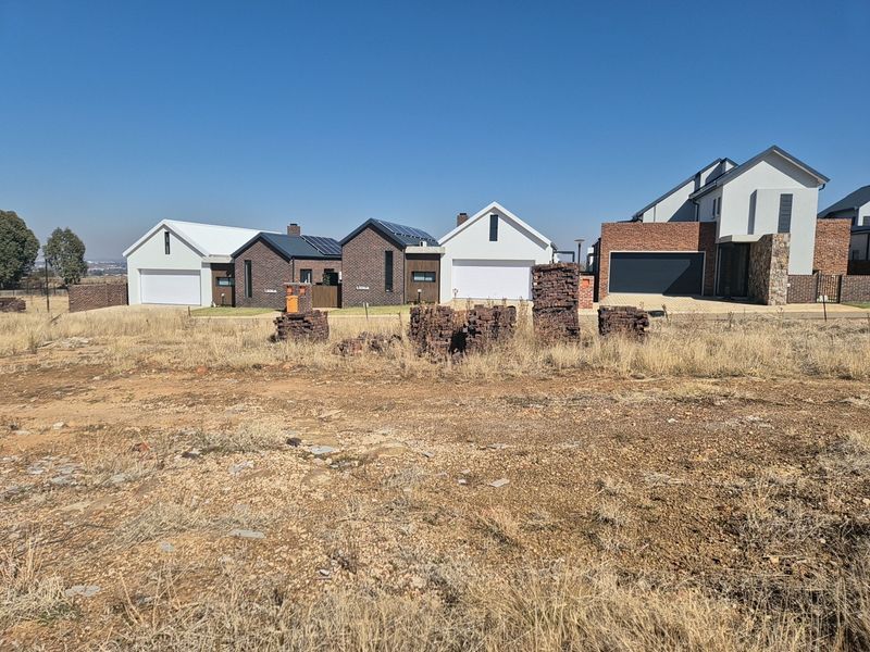 A view of a vacant residential plot with established modern homes in the background, showcasing the surrounding neighborhood development.