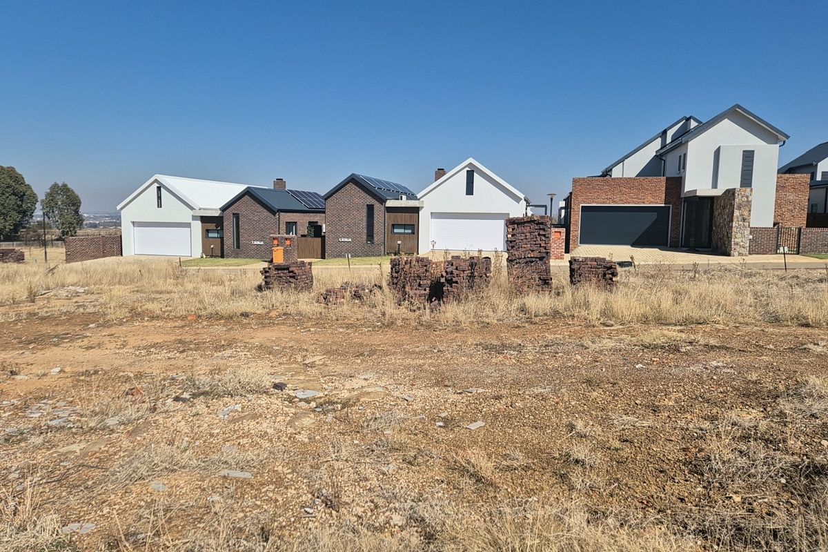 A view of a vacant residential plot with established modern homes in the background, showcasing the surrounding neighborhood development.