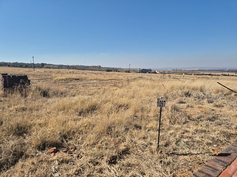 A vacant residential plot of land featuring dry grass, a boundary marker, and distant views of the surrounding estate.