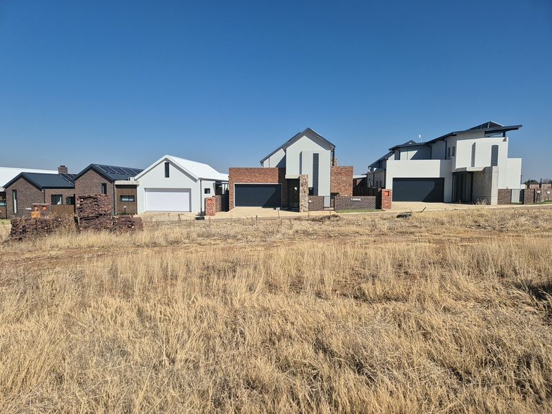 A view of modern residential homes with brick and rendered facades, garages, and solar panels, situated adjacent to an undeveloped vacant plot.