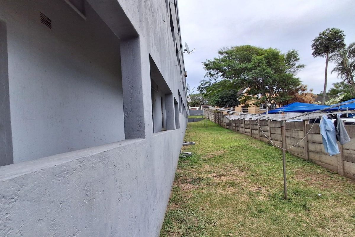 View of the apartment building's rear exterior, featuring a concrete facade, open walkway, and a grassed garden area enclosed by a boundary wall.