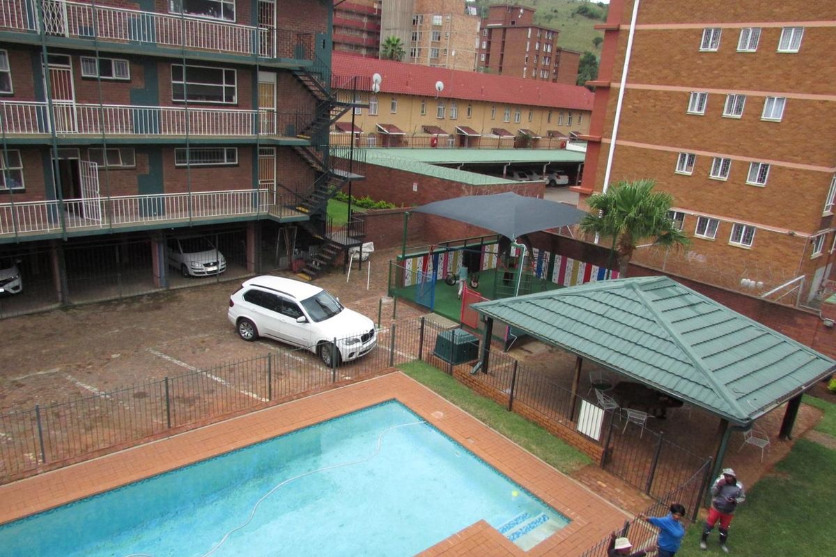Elevated view of the apartment complex communal area featuring a swimming pool, covered patio, and paved parking facilities.