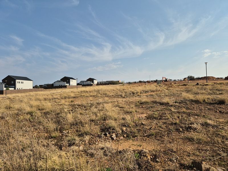 A view of a vacant residential plot featuring dry grass and surrounding neighborhood development.
