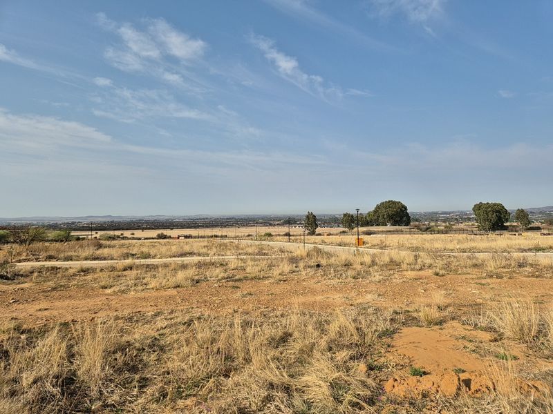 A wide-angle view of a vacant residential plot featuring dry grass, perimeter fencing, and distant landscape views.
