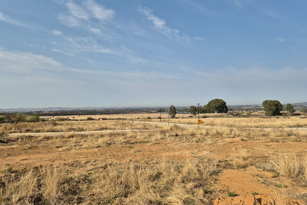 A wide-angle view of a vacant residential plot featuring dry grass, perimeter fencing, and distant landscape views.
