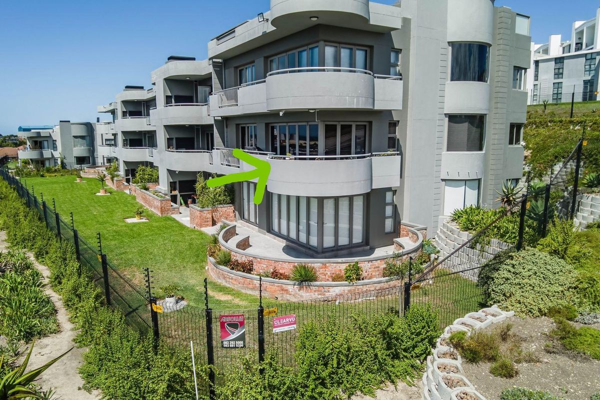 An elevated exterior view of a modern apartment complex featuring curved balconies, large glass sliding doors, and landscaped garden areas with perimeter fencing.