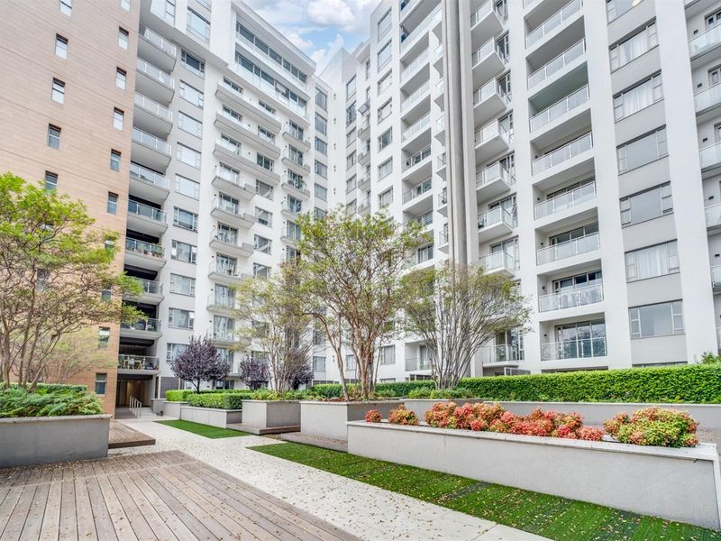 A well-maintained apartment complex courtyard featuring landscaped garden beds, raised concrete planters, and a timber deck walkway between residential building wings.