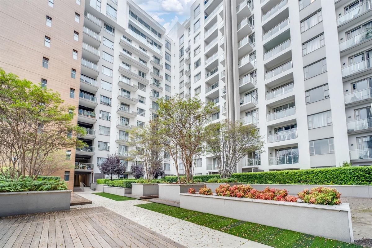 A well-maintained apartment complex courtyard featuring landscaped garden beds, raised concrete planters, and a timber deck walkway between residential building wings.