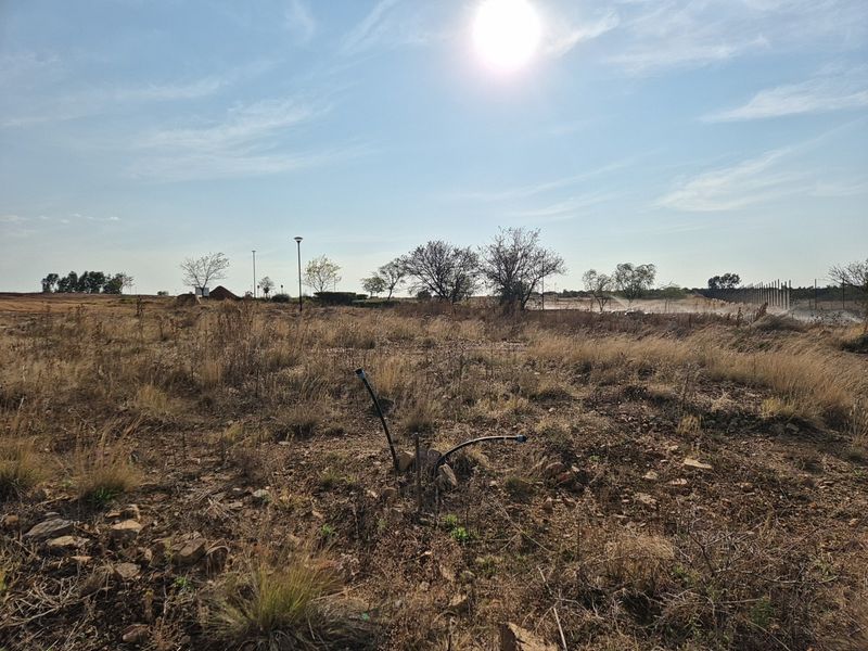 A view of a vacant residential plot featuring dry grass, scattered rocks, and visible utility connection points under a bright sky.